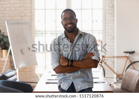 Smiling confident african american young businessman standing with folded hands, looking at camera. Portrait of happy handsome male manager satisfied with company career at modern creative office.