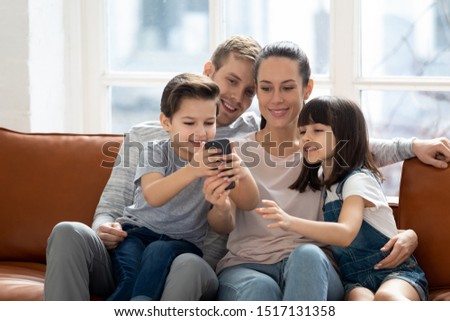 Excited family of young dad, smiling mommy, school daughter and little cute son sitting on couch, hugging, entertaining, using mobile phone, watching funny video, making purchases in shopping app.
