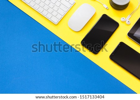 Top view of blue and yellow desk table with smartphone, mouse, keyboard, loudspeaker, charging head, laptop and earphone. Copy space, flat lay.