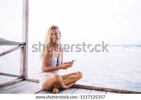 Positive beautiful blond female in white summer clothes smiling brightly and looking away while sitting on wooden pier and surfing internet in modern digital gadget on nature