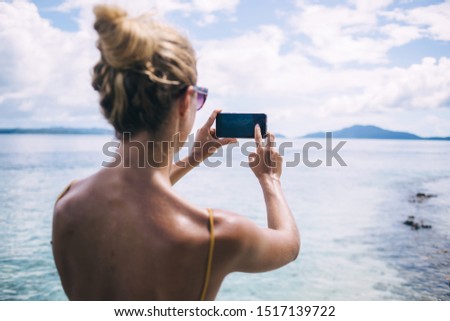 Back view of slim female in blur using smartphone and taking picture distant island in picturesque ocean landscape having vacation 