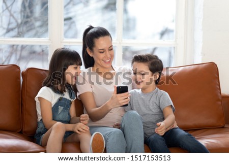 Happy young mommy sitting with children siblings, holding mobile phone in hands, showing funny video to kids, laughing, having fun together, using shopping application in living room at home.