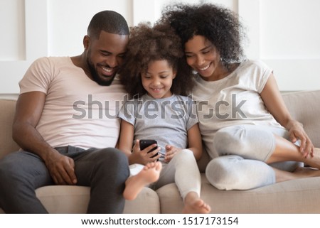Family looks at device screen african couple little daughter sitting on sofa kid girl holding gadget watching with parents cartoon browse internet together, parental control, have fun, new app concept