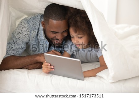 Close up african father little daughter lying in bed with tablet hiding under white blanket, have fun in internet, online apps, play games, watch cartoons, common mystery secrecy share secret concept