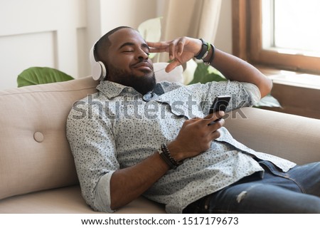 African calm guy hold smart phone fell asleep lying on couch listens music on headphones enjoy song or meditation audio course feels serenity inner harmony and balance, no stress mood, leisure concept Royalty-Free Stock Photo #1517179673