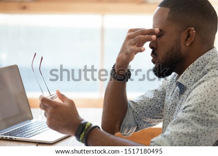 Close up side view african man sitting at desk near computer take break from study or work took off glasses rub nose bridge to reducing eye strain. Dry eyes or poor vision caused by pc overuse concept Royalty-Free Stock Photo #1517180495