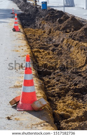 An emergency orange-and-white striped cone on asphalt near a roadwork, with a trench and pieces of brown clay
