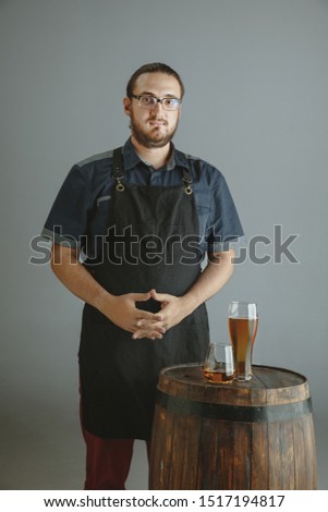 Confident young male brewer with self crafted beer in glass on wooden barrel on grey background. Man with hands crossed presented his products of brewing. Oktoberfest, drink, alcohol, industry.