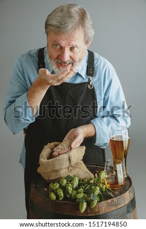 Confident senior man brewer with self crafted beer in glass on wooden barrel on grey background. Owner of factory presented his products, testing quality. Oktoberfest, drink, alcohol, industry.