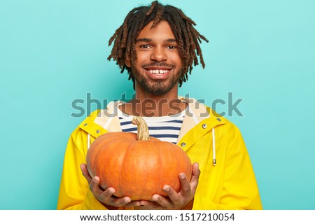 Cheerful dark skinned man in yellow raincoat boasts his autumn crop, holds pumkin, smiles pleasantly, poses over blue background, prepares for Halloween holiday. People, harvesting, healthy nutrition