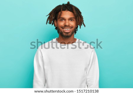 Dark skinned hipster with dreadlocks, smiles broadly, shows teeth, wears white casual jumper, satisfied ater spending weekend with friends, isolated over blue background. People, ethnicity concept