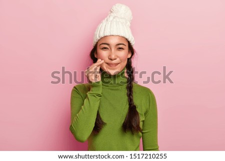 Pretty adorable mixed race girl makes korean heart gesture, has long hair combed in plaits, wears warm knitted hat and casual turtleneck, has natural beauty, isolated over pink studio background
