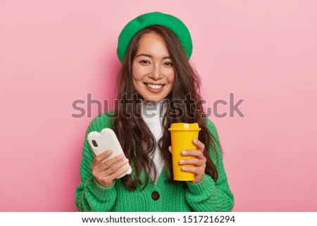 Portrait of positive brunette girl has coffee break after lectures, uses modern mobile phone for viewing pictures in social networks, sends notification to friend, wears stylish green beret, jumper