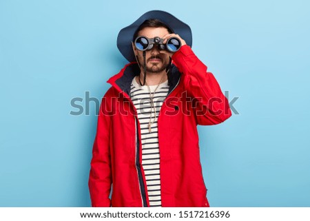 Male hunter looks through binoculars, wears hat and warm jacket, has adveturous journey, has active vacation during autumn, poses against blue background, watches something interesting discovers place