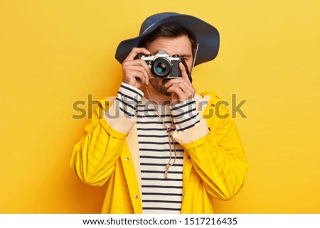 Say cheese. Young active male traveler takes photo with retro camera, dressed in hat, raincoat as travels during rainy day, poses against yellow background, has awesome trip, exlpores new places