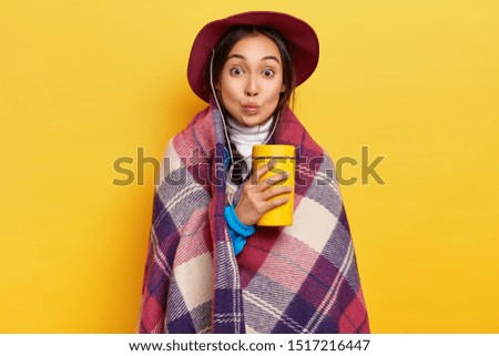 Lovely surprised female traveler has active camping rest, warms with hot beverage and checkered plaid, keeps lips folded, poses against yellow studio background. Asian girl with takeaway coffee