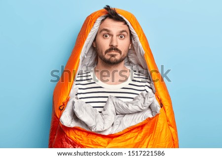 Photo of surprised bearded male camper poses in sleeping bag, wears striped jumper, spends weekend actively, poses over blue background, has expedition near rocky mountains. Active rest concept