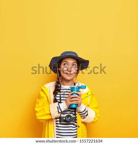Vertical image of lovely thoughtful girl in stylish headgear, protective raincoat, holds thermos with hot drink, carries retro camera for making photographes, creats content for social media channel