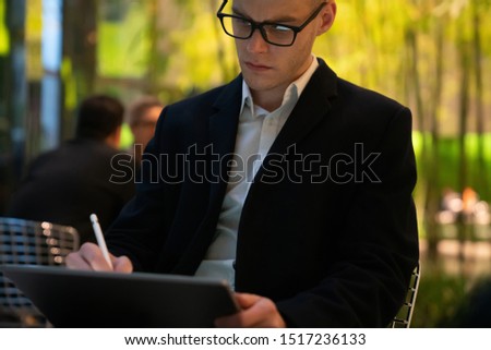 Crop serious employee in white shirt and black jacket with eyeglasses sitting and using tablet with pen on blurred background