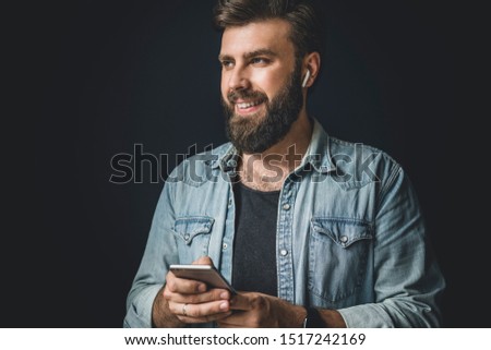 Smiling young man listening to favourite music on smartphone. Bearded hipster using wireless earphones. Portrait of male person in denim jacket with smartphone and headphones. Digital sound technology