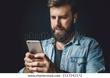 Bearded man in denim jacket using smartphone in everyday life. Modern lifestyle of digital specialists. Guy with wireless earphones listening to music online. Male person taking selfie on smartphone.