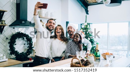 Joyful modern multiracial young men and women in Santa hats and casual clothes taking photo with smartphone while standing by table at decorated kitchen
