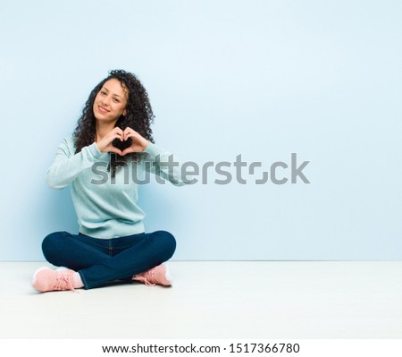 young pretty woman smiling and feeling happy, cute, romantic and in love, making heart shape with both hands sitting on floor