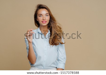 Pleased lovely woman with long hair, keeps hand raised, wears fashionable shirt, has clam face expression, smiles pleasantly, stands in studio against brown background. People and fashion concept
