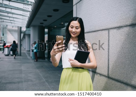 Joyful stylish young Asian woman in casual clothes with planner taking photo on smartphone while standing beside urban office building in daylight