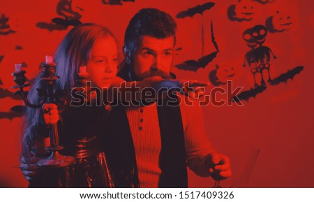 Girl and bearded man with suspicious faces on red background. Father and daughter with Halloween decor. Dad and kid in dress and scarf hold chandelier and point straight. Halloween night concept