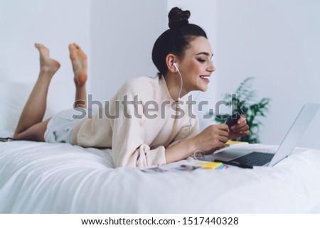 Side view of casual woman with creative hairstyle lying on bed using laptop with earphones enjoying and holding photo card