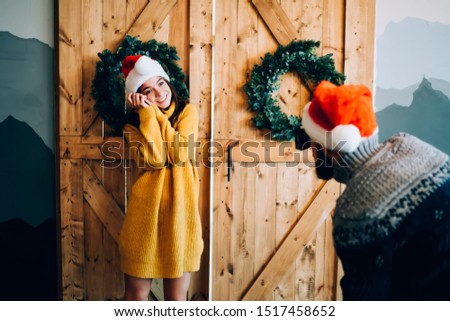 Beautiful cheerful young woman wearing warm winter sweater and red Santa hat posing for Christmas pictures and smiling at man with camera