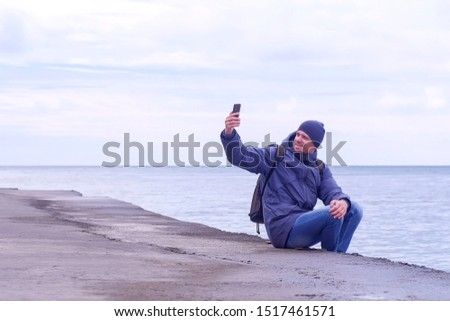 Tourist in warm clother hat and jacket takes a pictures on mobile phone on sea background. Man traveller makes selfie on smartphone aits on waterfront at sea in winter. Overcast rainy day at seaside.