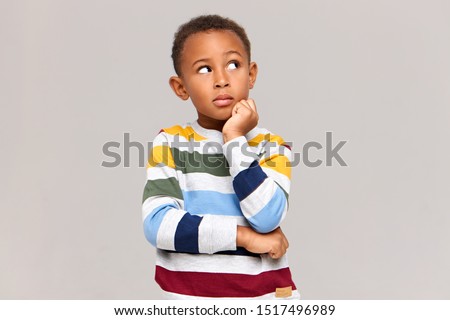 Studio picture of cute dark skinned nine year old boy keeping hand under his chin and looking up with thoughtful pensive facial expression, having curious look, trying to remember something