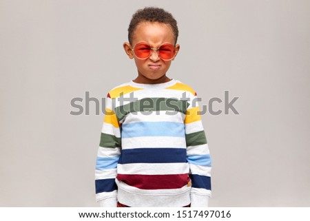 Picture of stubborn Afro American kid in stylish sweater and pink shades grimacing, screwing up face, does not want to do homework or go to school. Human facial expressions, emotions and feelings