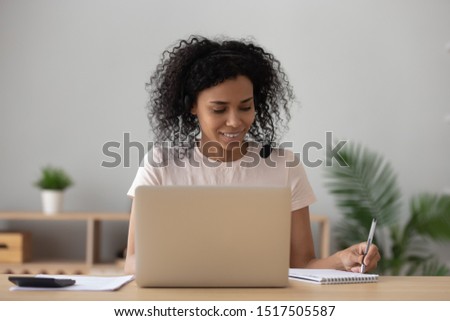 African focused woman sitting at desk wearing headset hearing online webinar lesson making notes in writing pad, 30 female student studying via internet using video conference call, e-learning concept Royalty-Free Stock Photo #1517505587