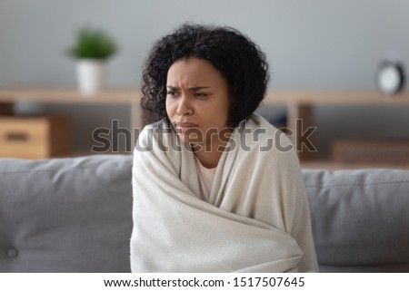 African unhappy woman sit on couch covered with plaid try to warming up in cold flat without central heating, system not working, girl feels unhealthy has common cold viral infectious disease concept Royalty-Free Stock Photo #1517507645