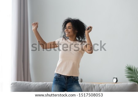 African woman holds smartphone enjoy favourite song listens tracklist in living room at home, girl dancing alone spend free time on weekend raise hands smiles moving, music admirer, hobby mood concept Royalty-Free Stock Photo #1517508053