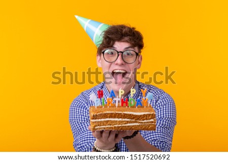 Crazy cheerful young man in paper congratulatory hat holding cakes happy birthday standing on a yellow background. Jubilee congratulations concept.