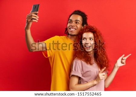 Happy lovely mixed race couple has stroll together, stand next to each other, pose for making selfie, being in good mood. Diverse friends take picture of themselves, red wall. Tourists go sightseeing
