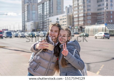 Two joyful attractive girls taking selfie while standing together at sidewalk and showing peace gesture outdoor