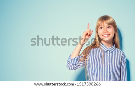 Beauty funny blonde teenage girl has an idea, pointing finger, showing empty copy space for text, blue background. Happy girl presenting point. School girl Proposing product. Advertisement gesture