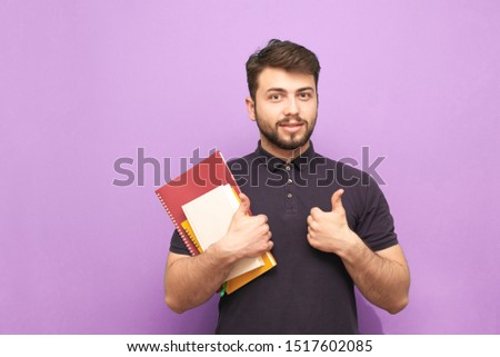 Portrait of a bearded male student standing with books and exercise books in his hands, smiling and pointing his thumb at the camera. Happy guy with books posing at camera.