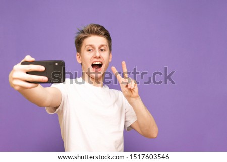  positive boy takes a selfie,wearing a white Tshirt, standing on a purple background and posing on the camera.Happy young man makes selfie. Shows a sign of peace in the camera of a smartphone