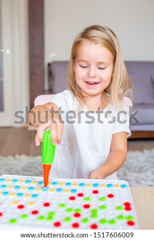 Happy pretty little blonde girl sitting at a table at home playing with a toy screwdriver and multicolor screws. Early education.