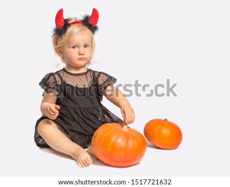 Emotional little baby girl in black dress and devil horns playing with pumpkins for Halloween celebration over white background.