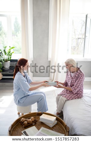 Nurse bringing book. Aged grey-haired woman feeling thankful to nurse bringing book