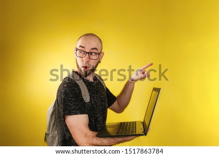 Advertising and business. A bald man in a black t-shirt and glasses, with a backpack, holding a laptop and pointing in surprise at something with his finger. Yellow background. Copy space
