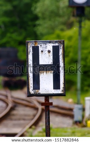 Vintage Railway enamel metal plate with a big “H“ letter black on white. Rusty and wheatherd surface with track, switch and signal in the background. 