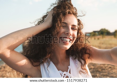 Photo of beautiful happy woman laughing and taking selfie photo while walking at seashore during sunny day